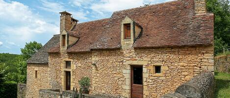 A stone house with a tiled roof, two chimneys, and a small window.