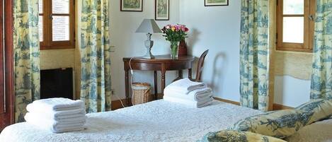 Quarto com cama, almofadas, mesa de cabeceira com abajur, vaso com flores e quadros na parede.