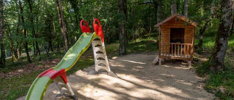 A playground with a slide and swings, a wooden playhouse, and a sandy area surrounded by trees.