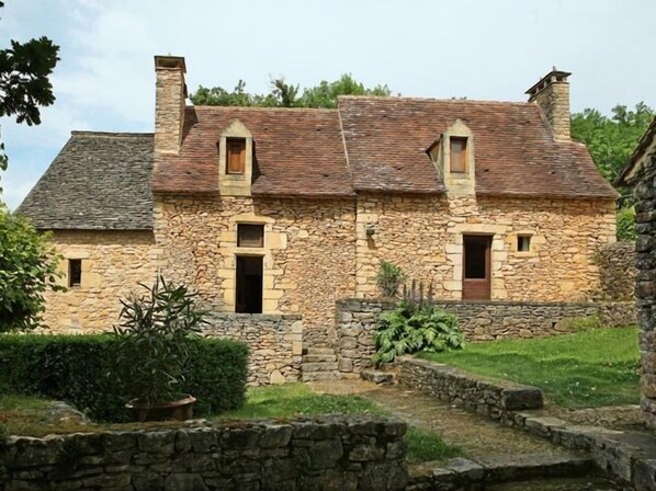 A stone house with a sloped roof, multiple chimneys, and a small garden area with a stone wall and potted plants. - Chambre D'hôtes La Rossillonie (Beynac-et-Cazenac)
