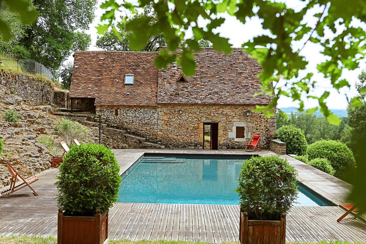 A stone house with a swimming pool, wooden deck, and red chairs.