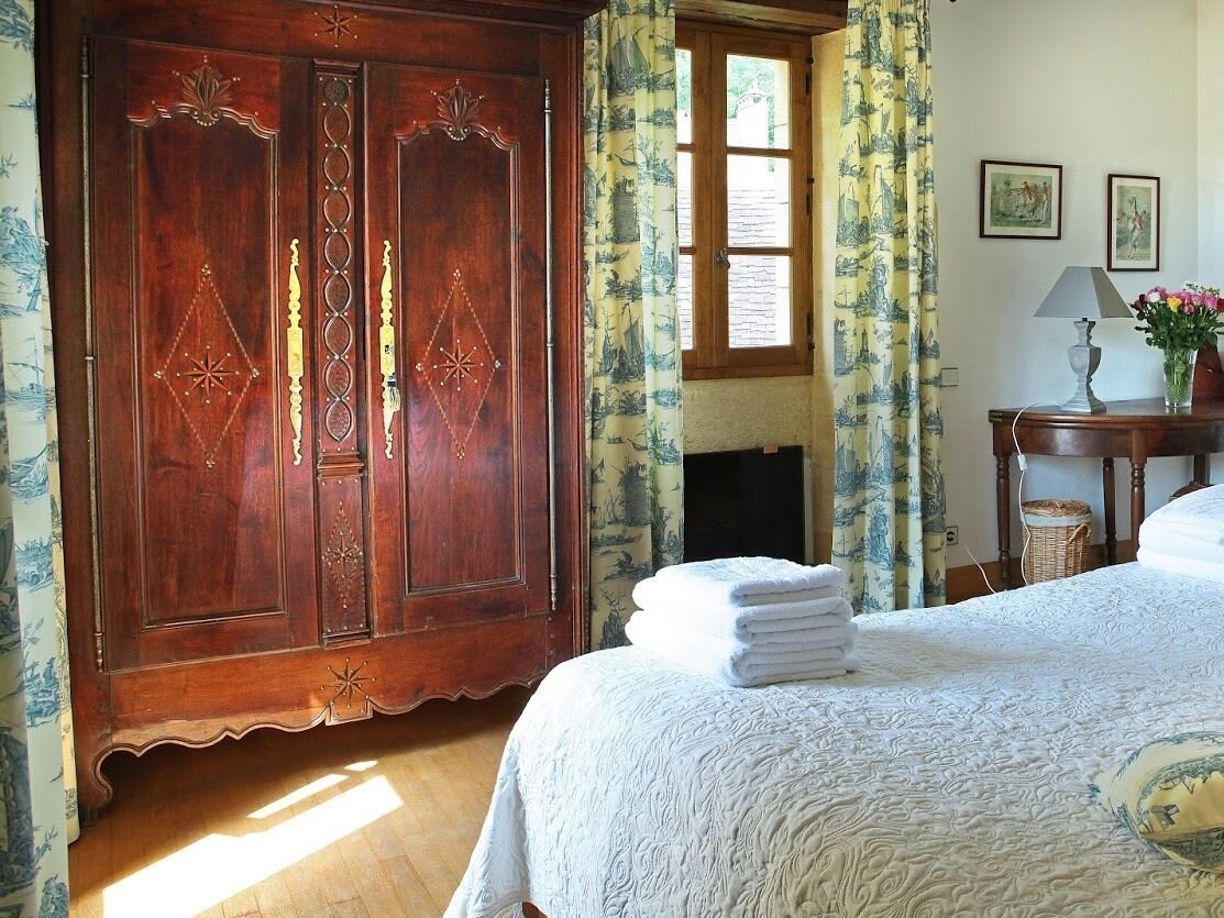 A bedroom with a large wooden wardrobe, a bed with a white bedspread, a window with curtains, a lamp on a bedside table, and a vase with flowers.
