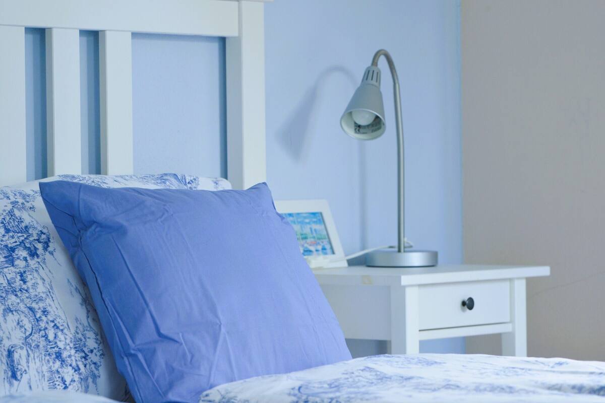 Een opgemaakt bed met blauw beddengoed, een bedlampje en een klein nachtkastje met een fotolijstje.