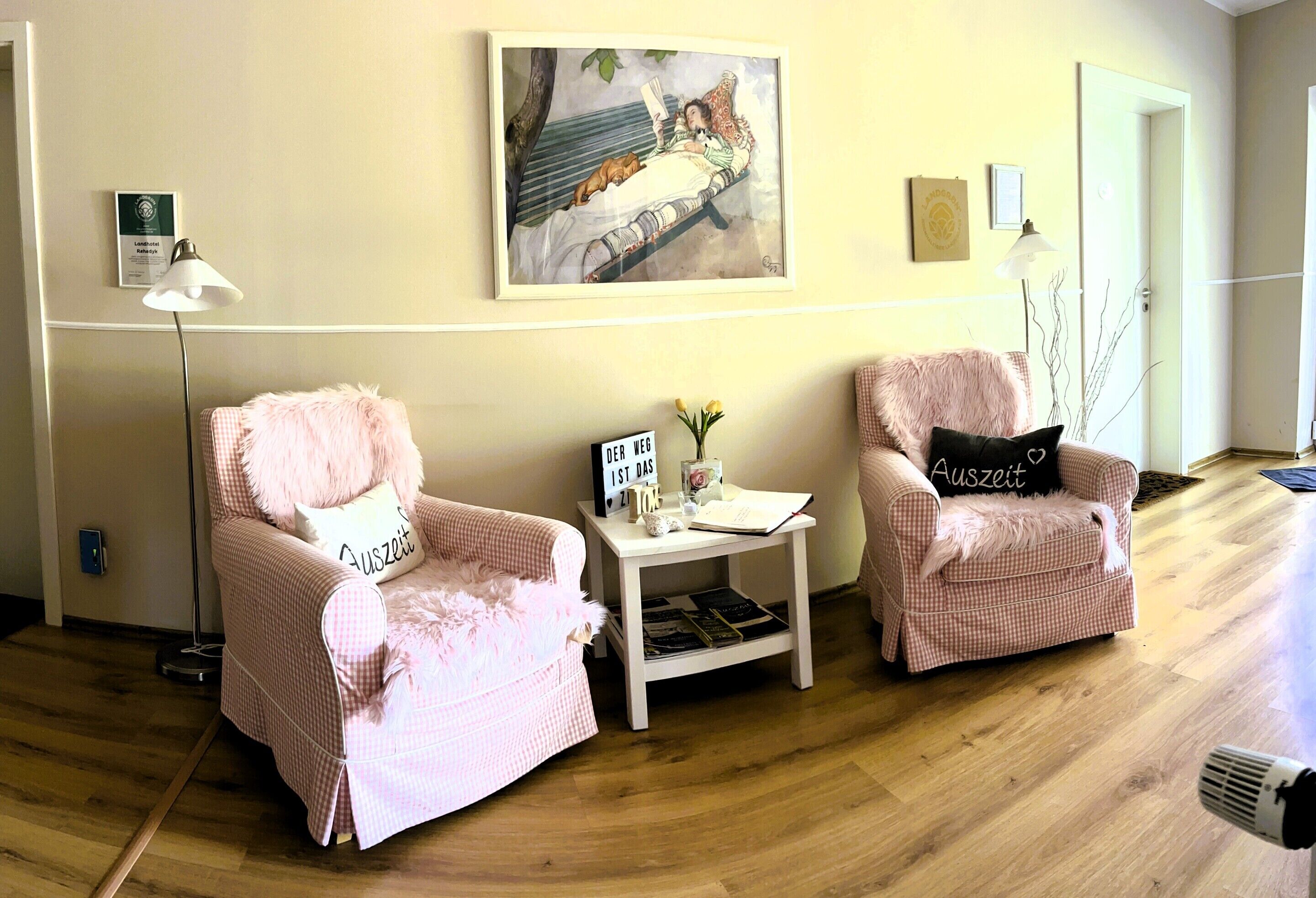 A room with two pink armchairs, a small white side table with a sign, a floor lamp, and a painting on the wall.
