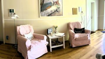 A room with two pink armchairs, a small white side table with a sign, a floor lamp, and a painting on the wall.