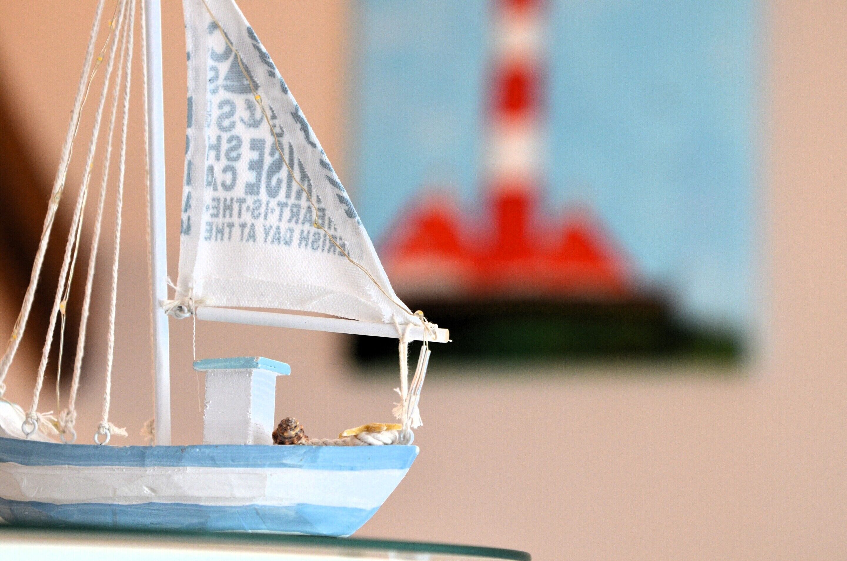 A model sailboat with a white and blue color scheme, a small figure inside, and a blurred background with a red and white structure.