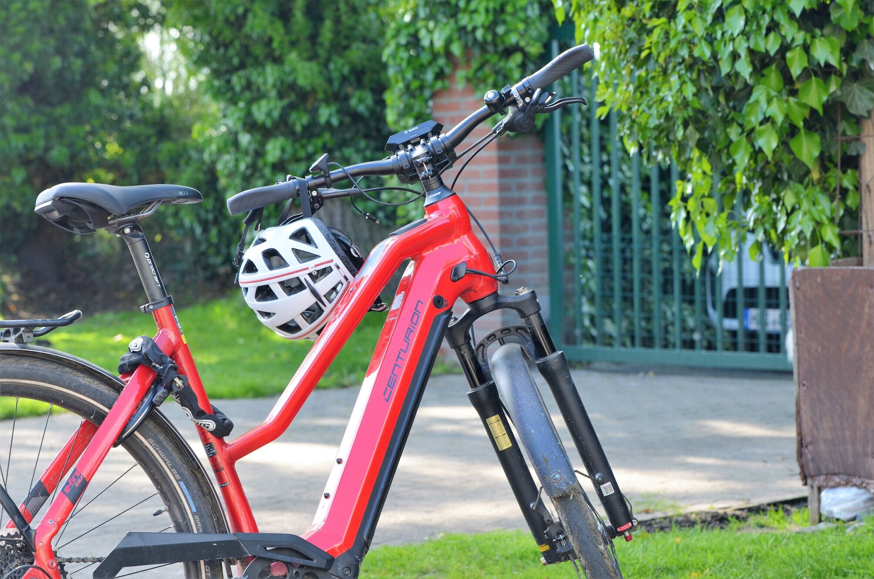 A red electric bicycle with a white helmet attached to the handlebars, parked on a sidewalk with a green fence and trees in the background.