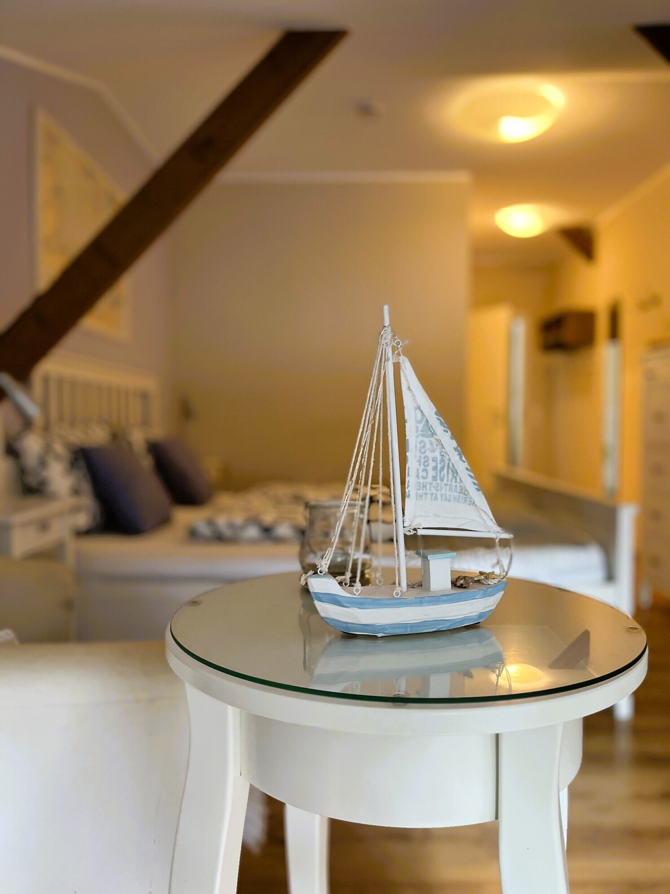 A model sailboat on a glass table in a room with a staircase and a bed in the background.