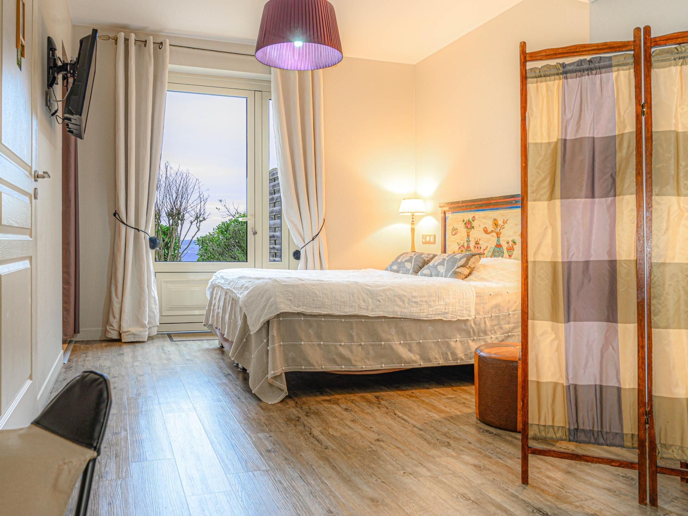 A bedroom with a bed, a desk, a chair, a window with curtains, a lamp, and a view of the outdoors.
