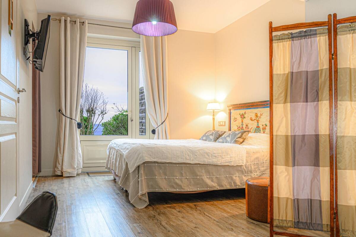 A bedroom with a bed, a desk, a chair, a window with curtains, a lamp, and a view of the outdoors.