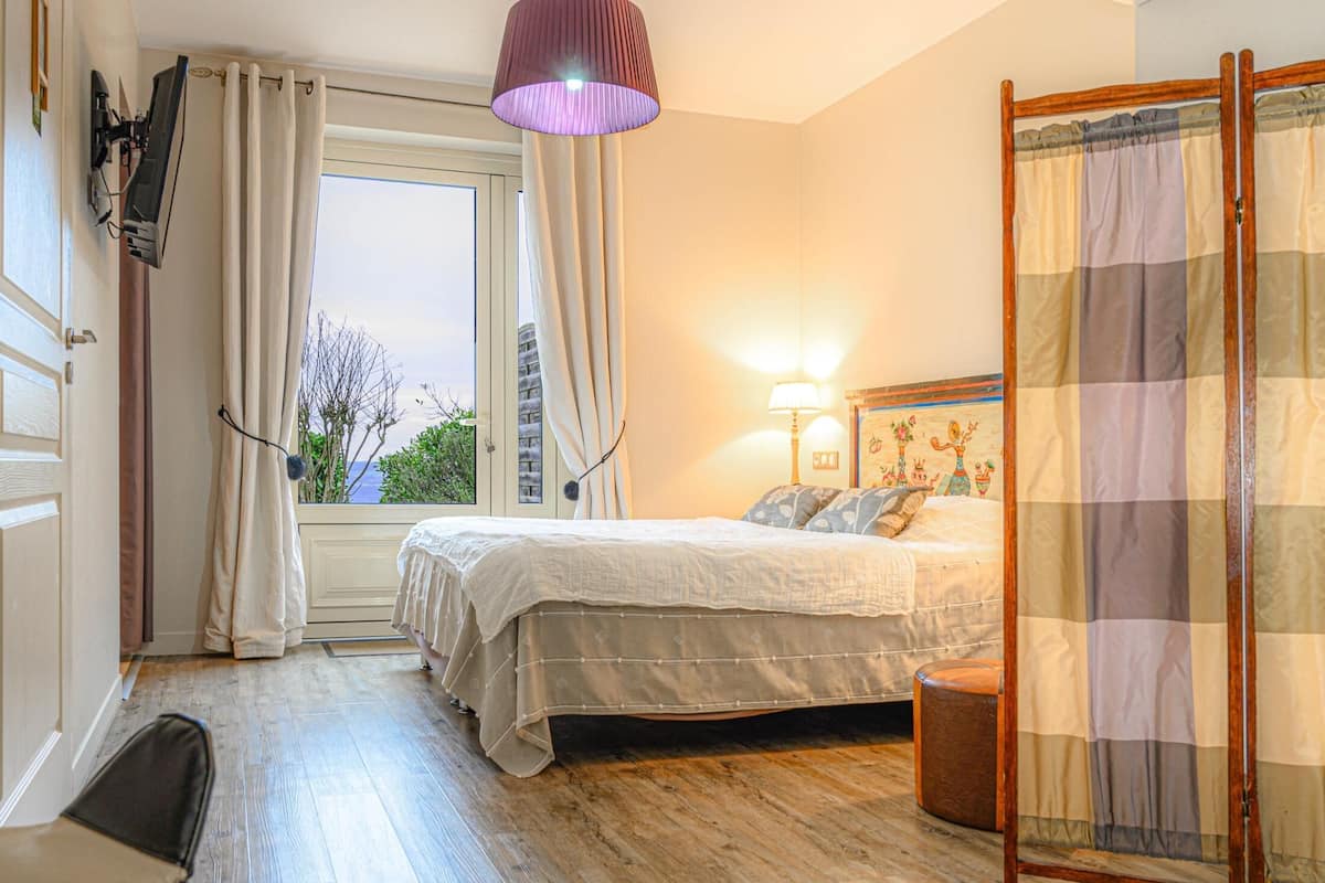 A bedroom with a bed, a desk, a chair, a window with curtains, a lamp, and a view of the outdoors.