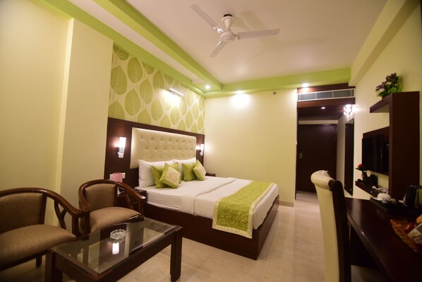 A hotel room with a bed, two chairs, a glass table, a ceiling fan, and a TV.
