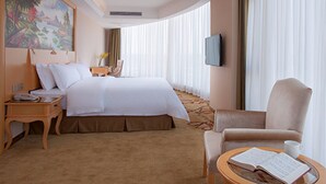 A hotel room with a large bed, a desk with a telephone, a chair, a round table with a book and a cup, and a painting on the wall.