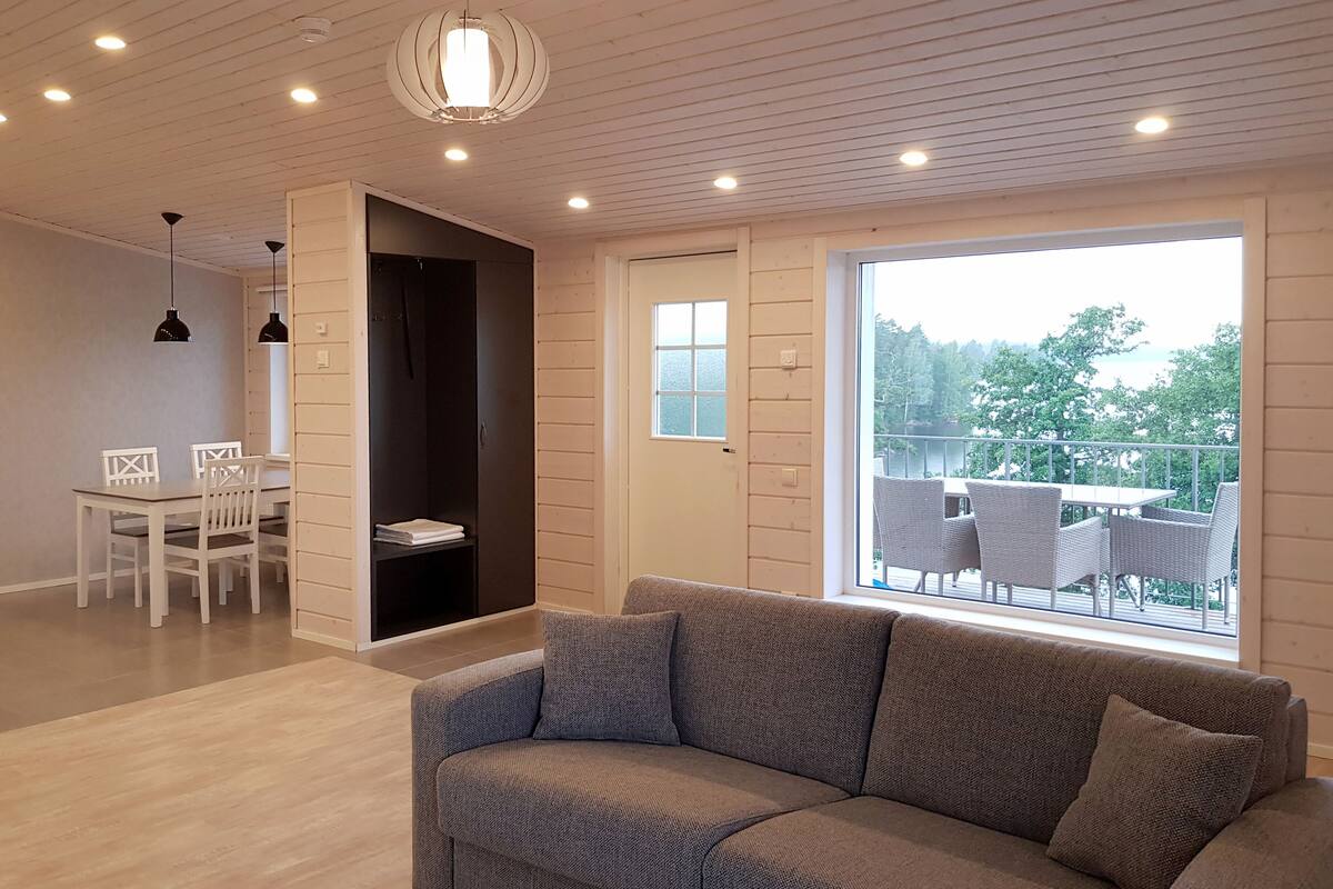 Een moderne woonkamer met een grijze bank, een eettafel met stoelen en een groot raam met uitzicht op de omgeving.