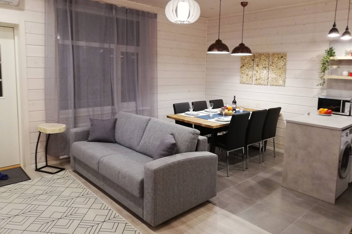 Een moderne woonkamer met een grijze bank, een eettafel met stoelen en een keukenhoek met een wasmachine.