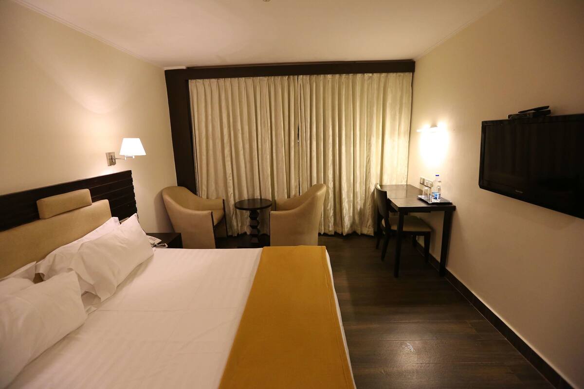 Habitación de hotel con cama, escritorio, silla, televisor y ventana con cortinas.