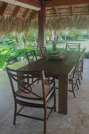 A wooden table with chairs in a covered outdoor area with a thatched roof, surrounded by greenery. - Luxury Villas at Tortuga Bay (Punta Cana)