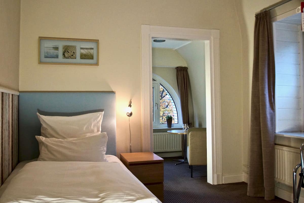 Ein Hotelzimmer mit einem Bett, einem Schreibtisch und einem Sessel. Es gibt ein Fenster mit Vorhängen, eine Heizung und ein Bild an der Wand.
