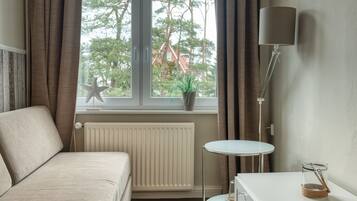 A bedroom with a bed, a small table, a lamp, and a view of trees and a house through a window.