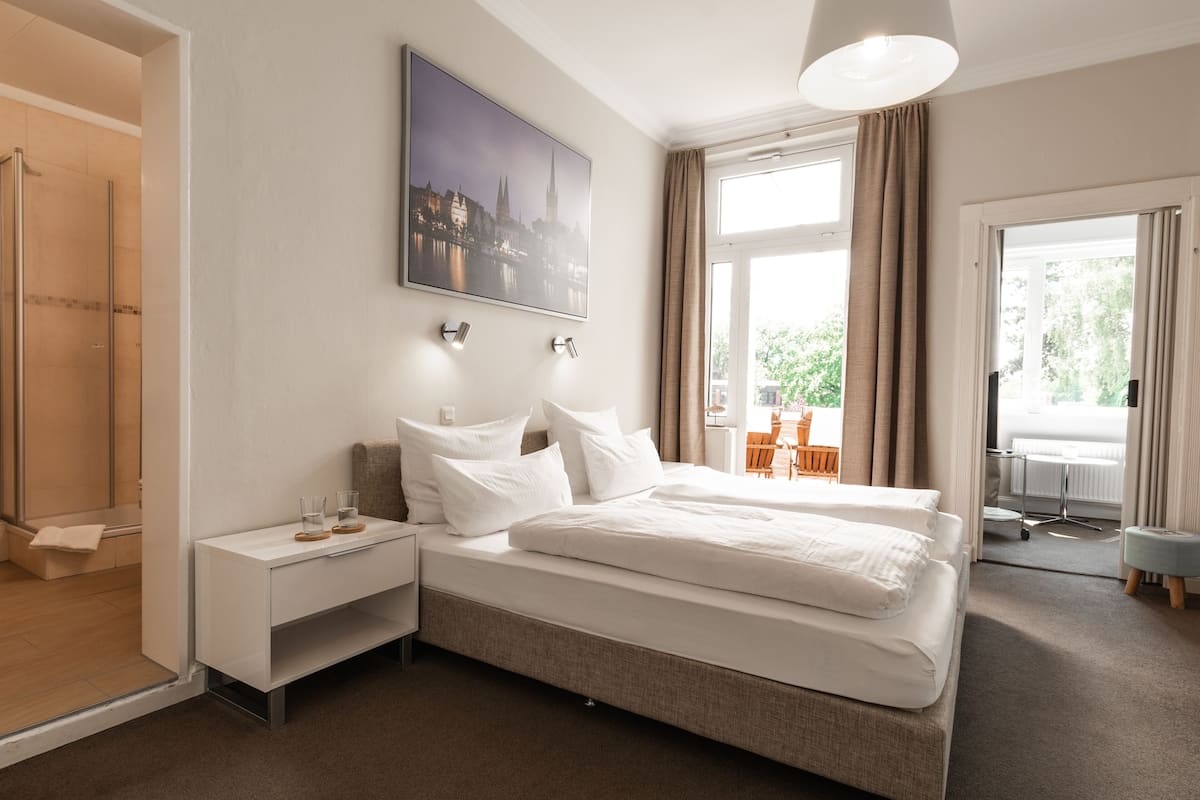 Ein Hotelzimmer mit einem großen Bett, einem Nachttisch mit Gläsern, einem Stadtbild als Gemälde und Blick ins Freie.