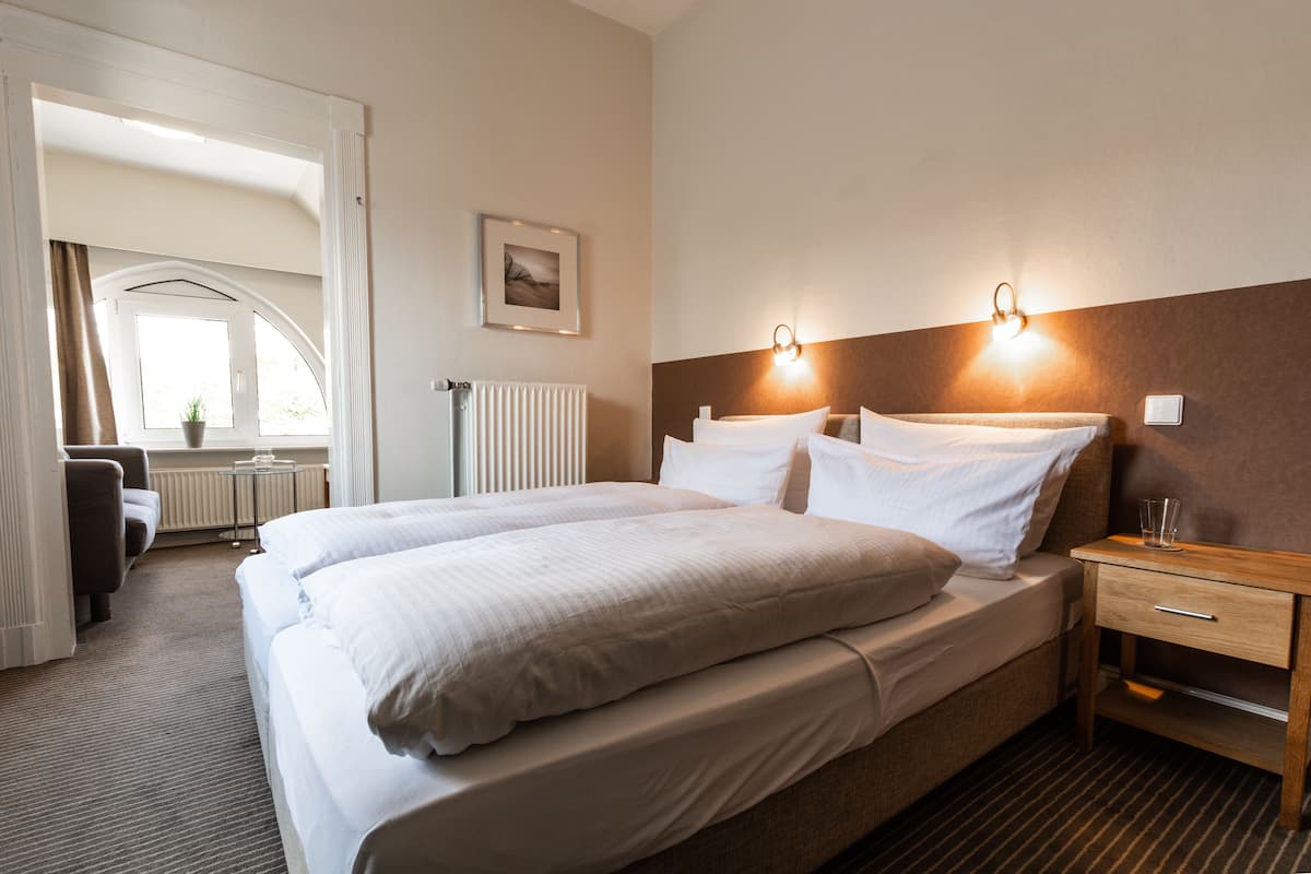 Ein Hotelzimmer mit Doppelbett, Nachttisch mit Glas, Wandleuchte, Heizkörper und Blick ins Freie durch ein Fenster.