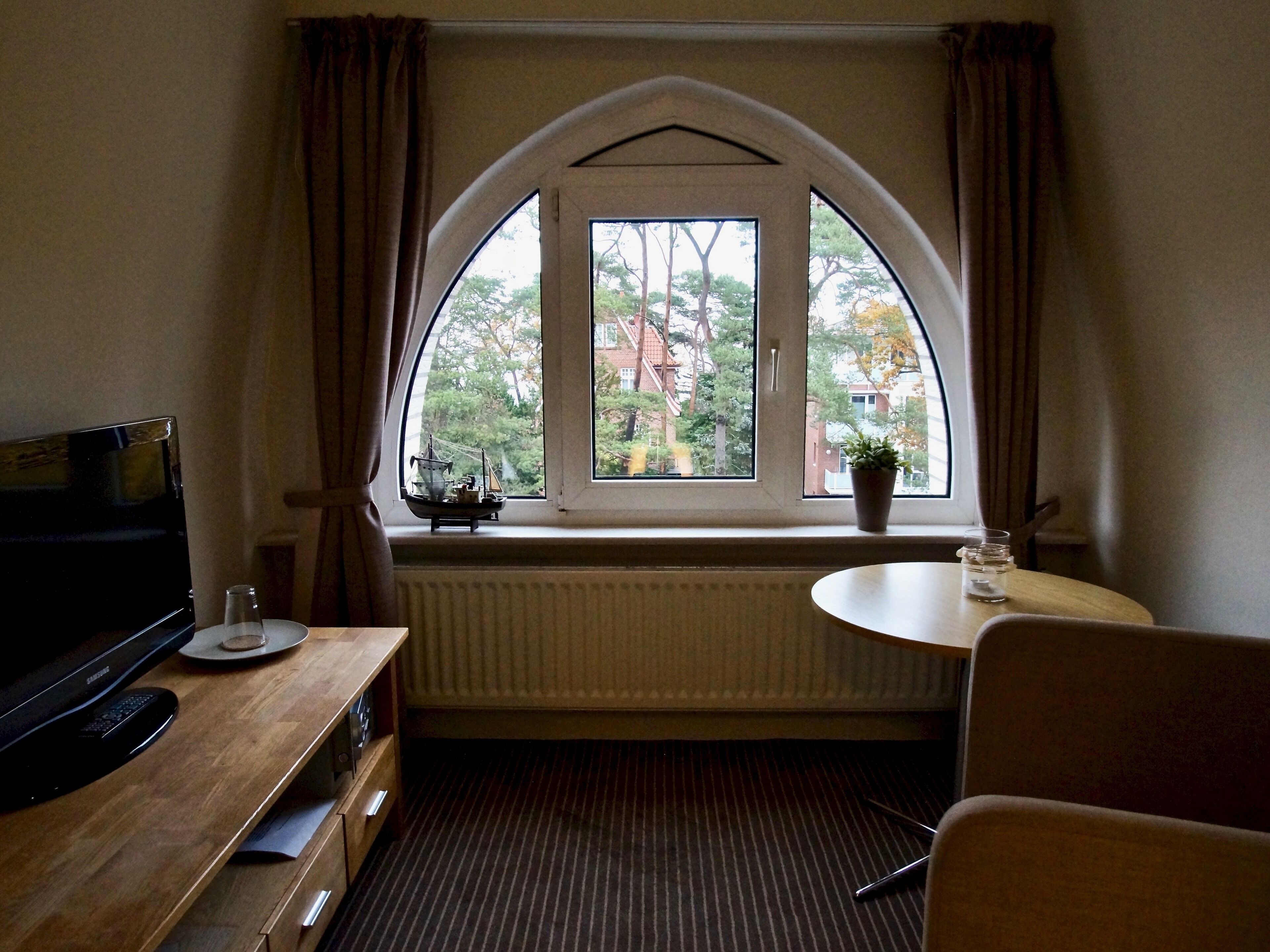 Une pièce dotée d’une grande fenêtre en arc de cercle, d’un bureau en bois avec un téléviseur, d’une petite table avec un verre et d’un radiateur.