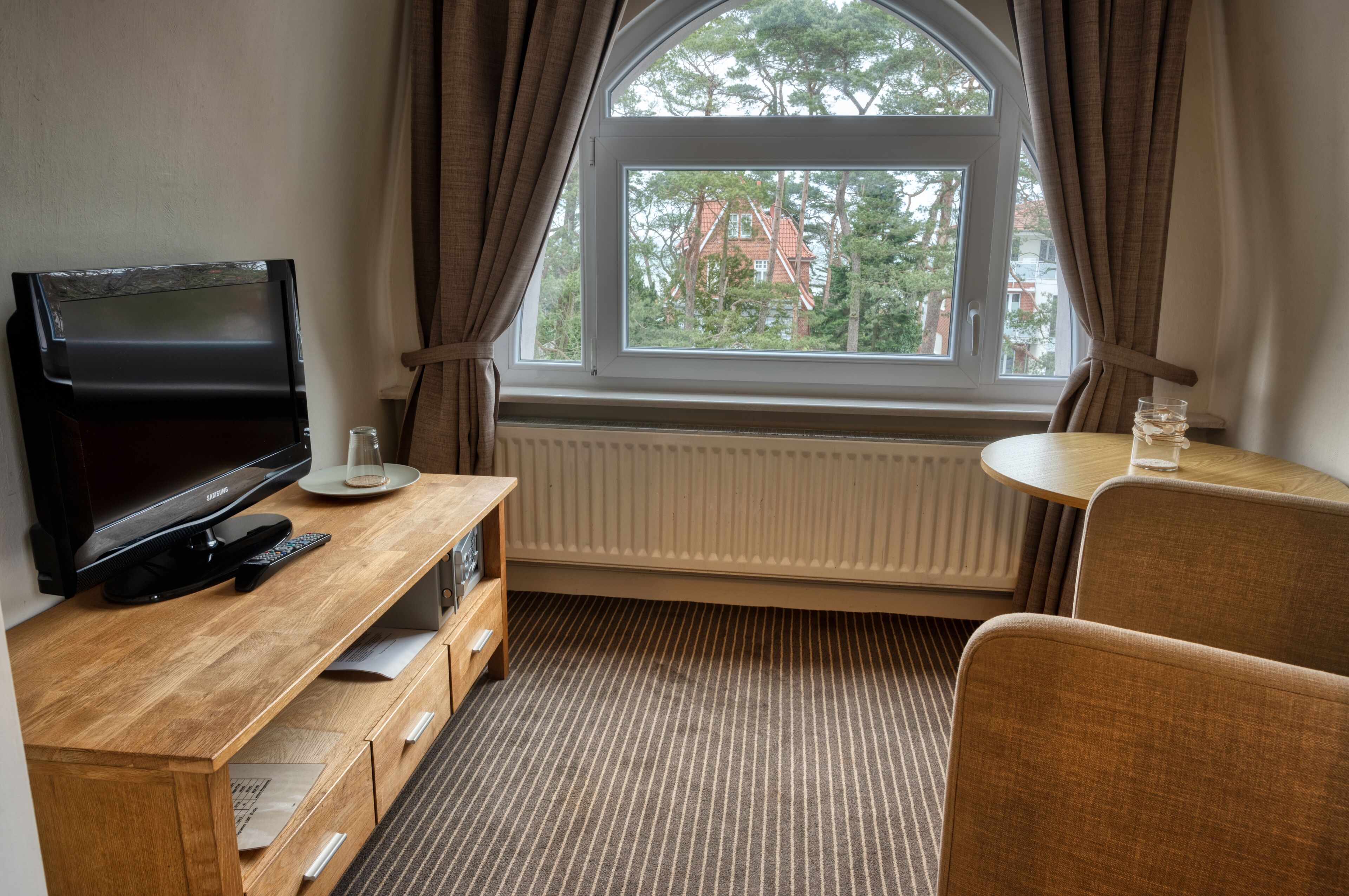 A hotel room with a TV, a small table, a chair, a radiator, and a window with curtains.