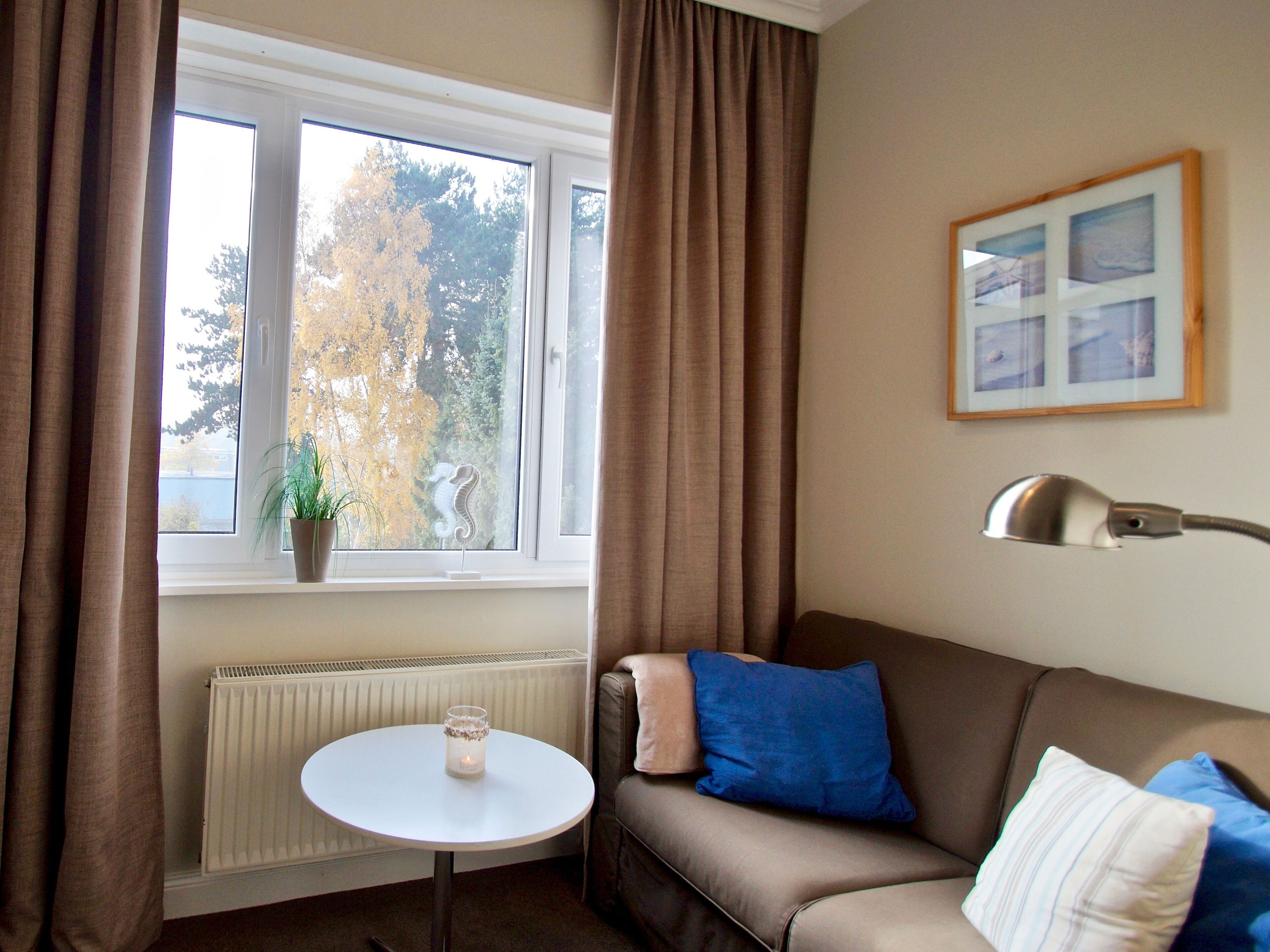 A room with a window, a radiator, a small table with a candle, a sofa with cushions, and a lamp on the wall.