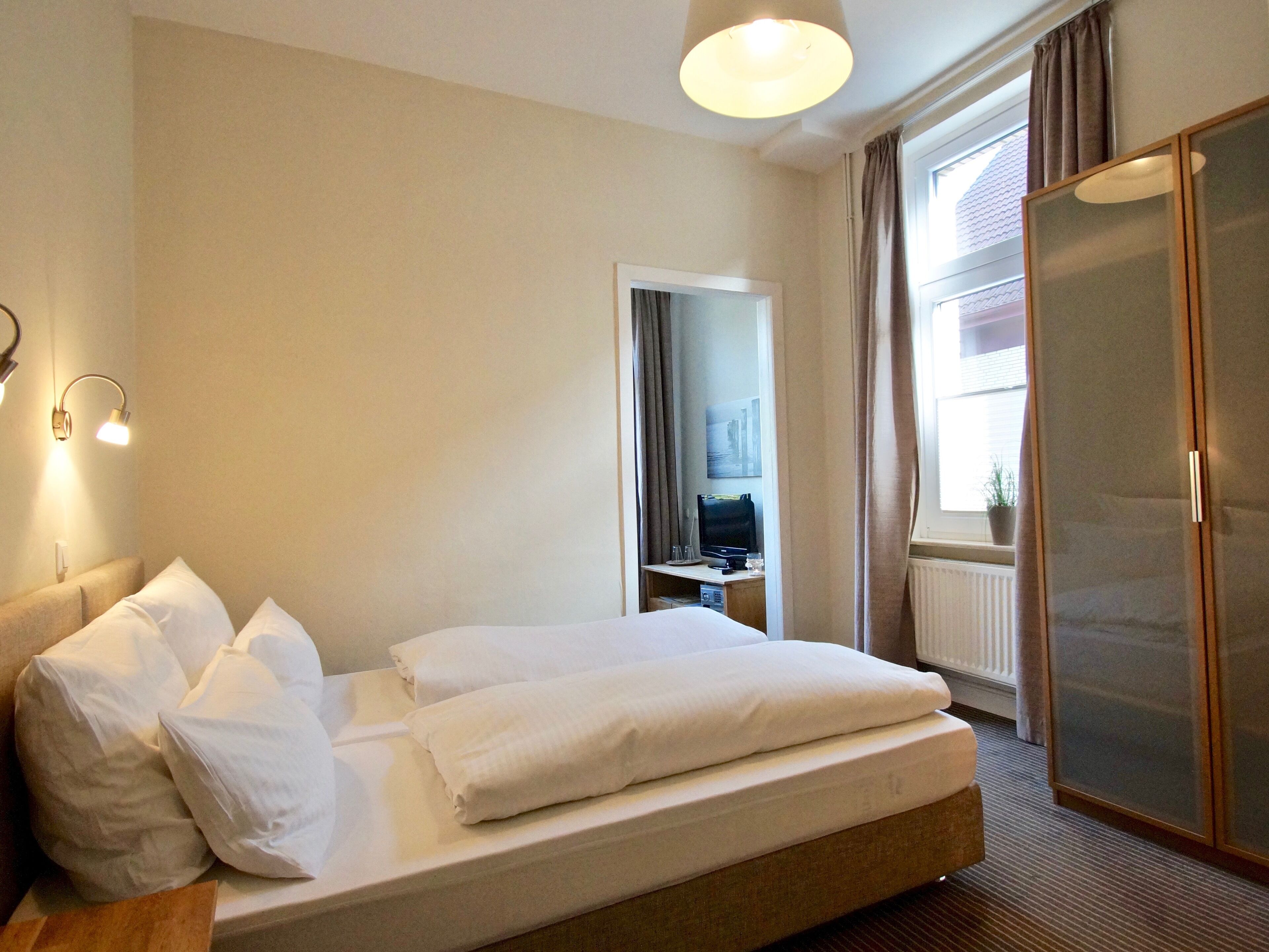 A hotel room with a bed, a desk with a computer, a wardrobe, and a window with curtains.