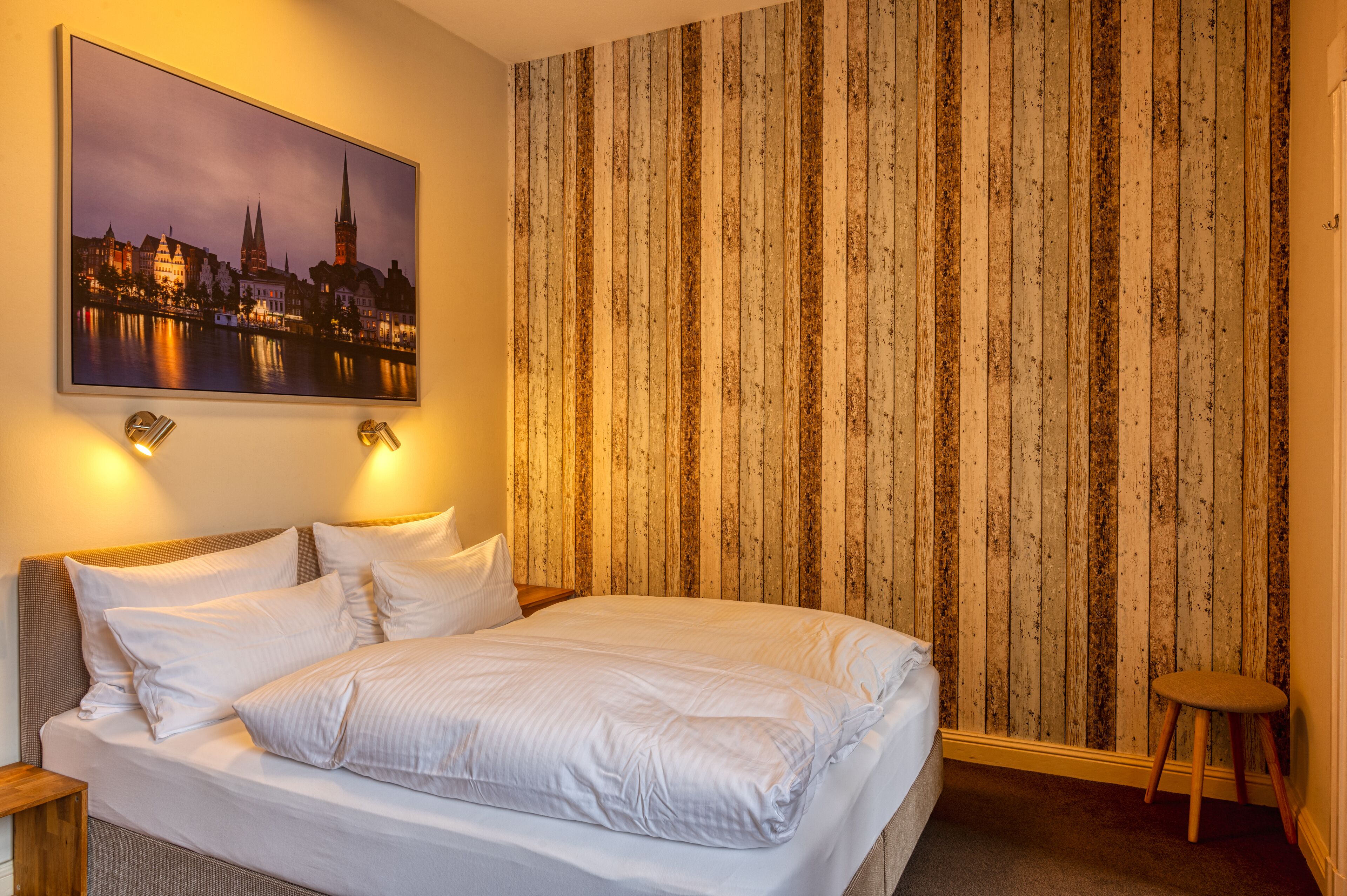 A neatly made bed with white linens, a cityscape painting above it, and a wooden striped wall.