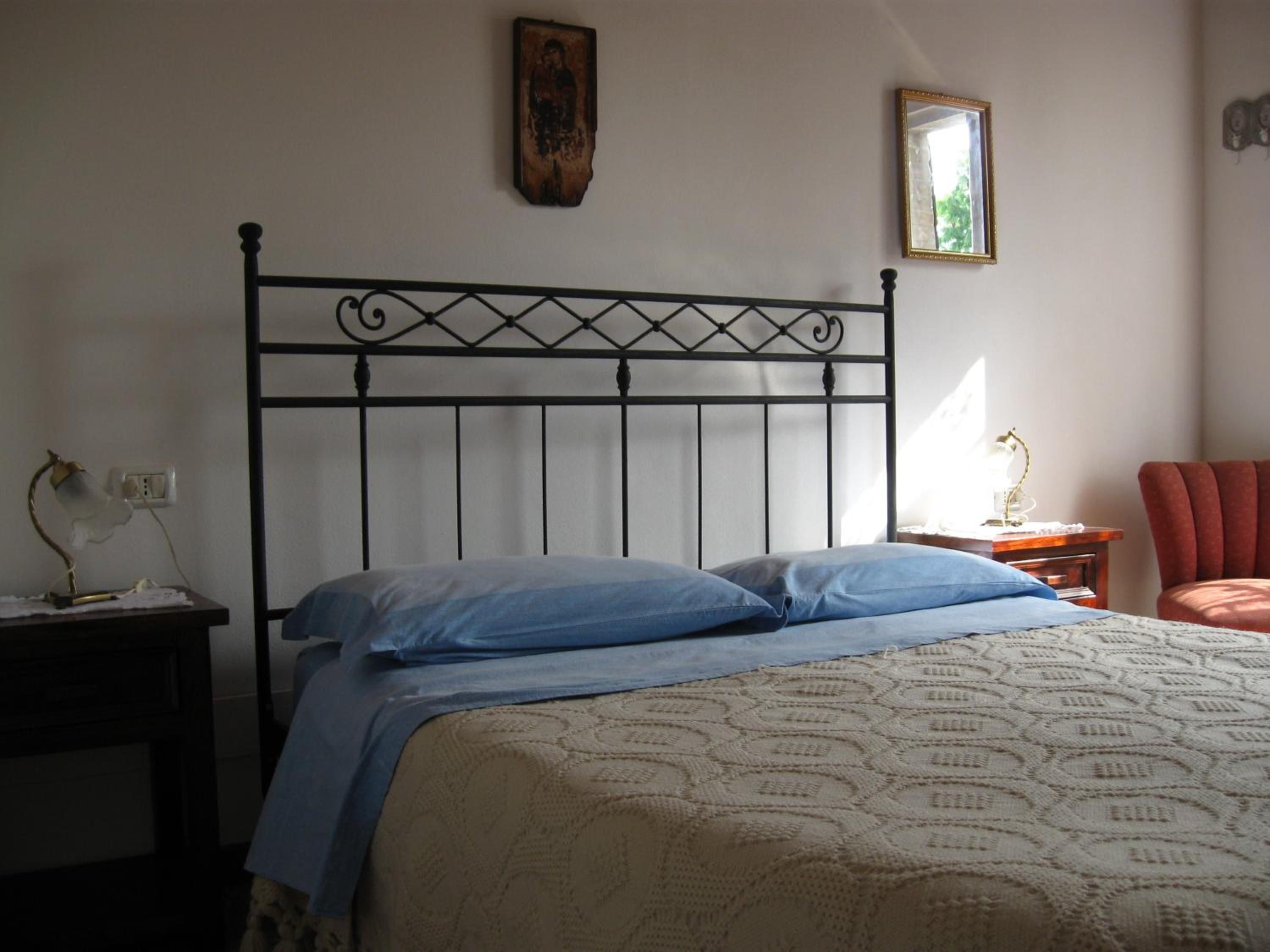 Un dormitorio con cabecera de metal, cama con ropa de cama azul, mesita de noche con lámpara y una silla roja.