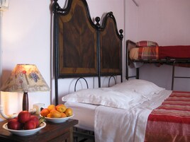 Un letto singolo con lenzuolo bianco e coperta rossa, un comodino con lampada e una ciotola di frutta, e un quadro appeso al muro.