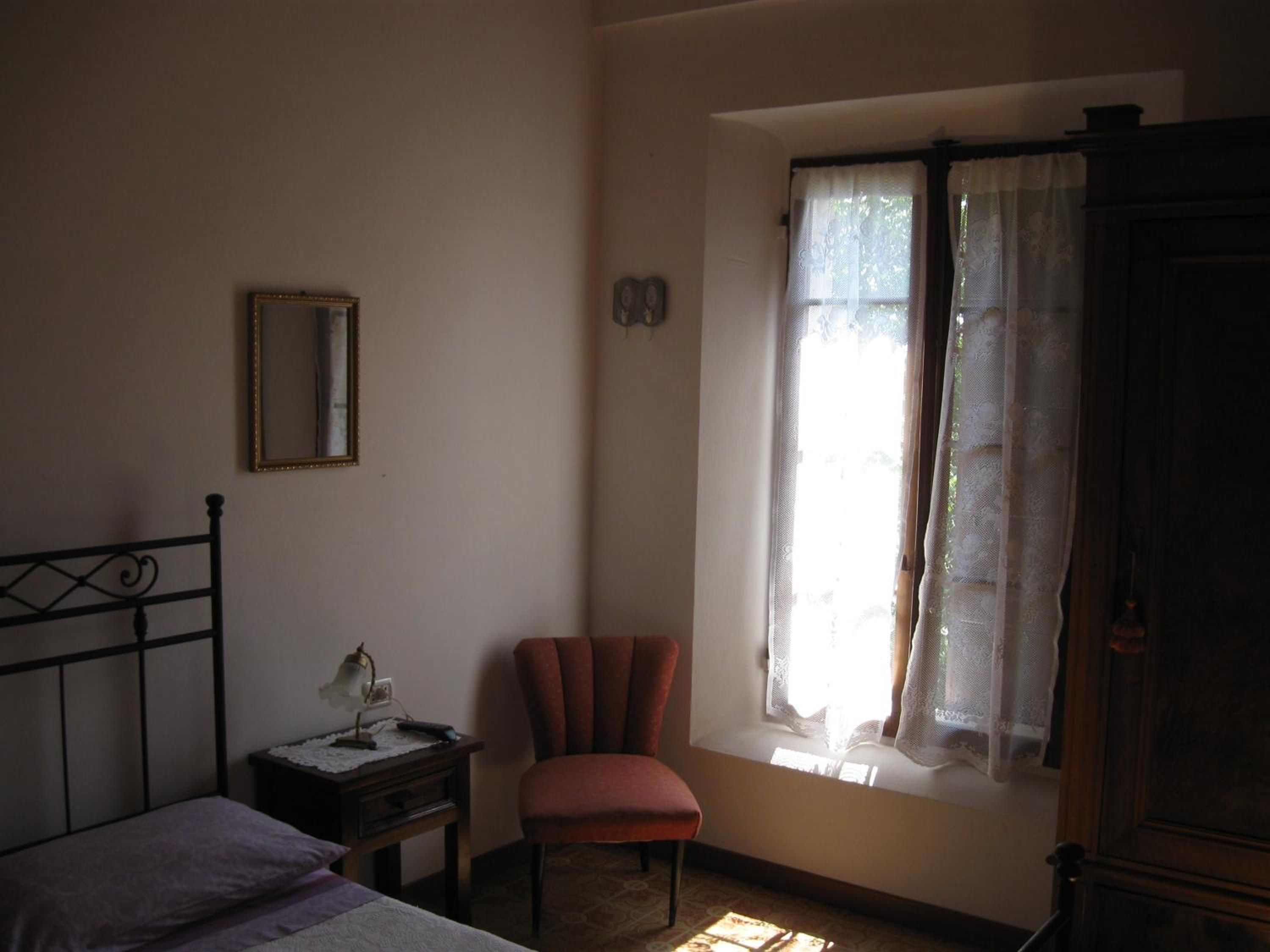 Un dormitorio con cama, una silla, una mesita de noche, una lámpara, una ventana con cortinas y un espejo en la pared.