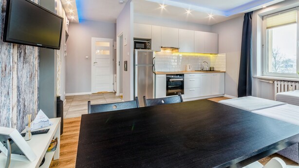 A modern kitchen with white cabinets, a built-in oven, and a microwave. There is a dining area with a black table and chairs.
