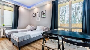 A room with two single beds, a dining table, and chairs. There are framed pictures on the wall and a window with curtains.