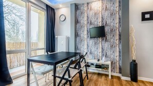 A modern room with a wooden floor, a dining table with chairs, a TV mounted on a textured wall, and a window with curtains.
