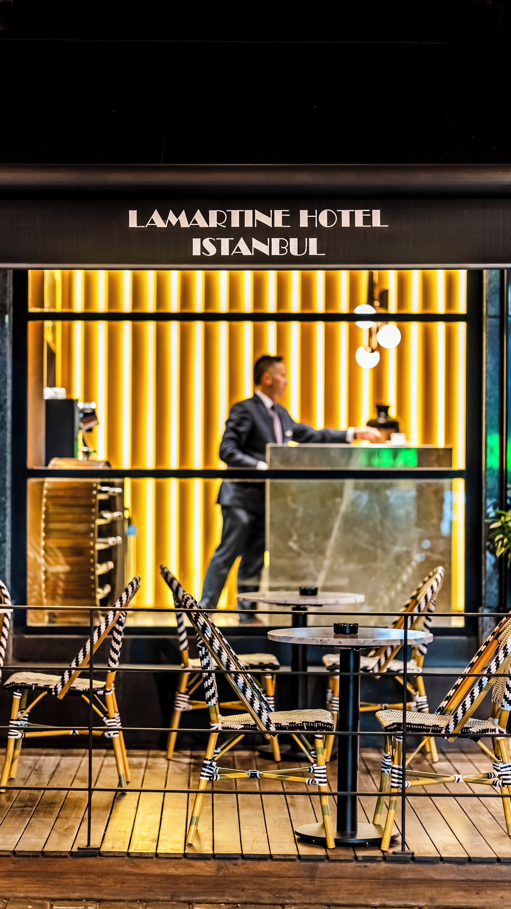 Et moderne interiør fra Hotel Lamartine i Istanbul, med et receptionsområde, en mand ved skranken, en glasvæg og unikt designede stole.