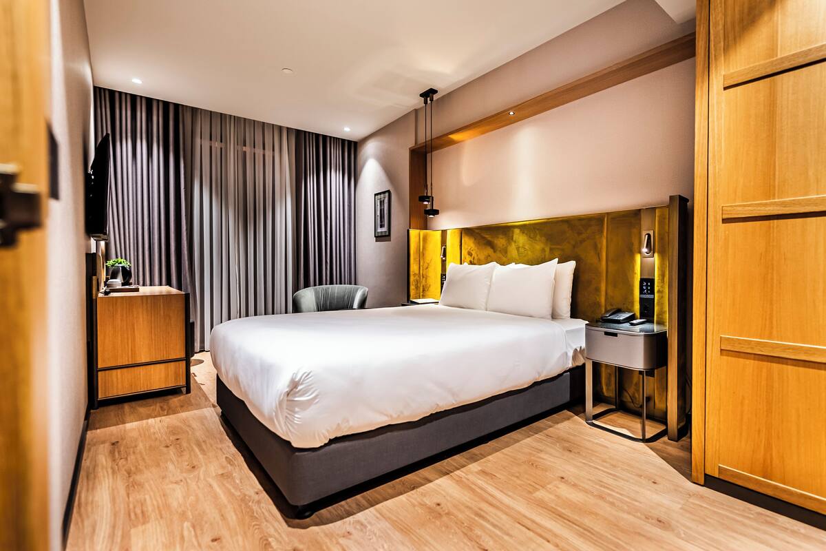 A modern hotel room with a large bed, a wooden wardrobe, a desk with a lamp, and a chair.