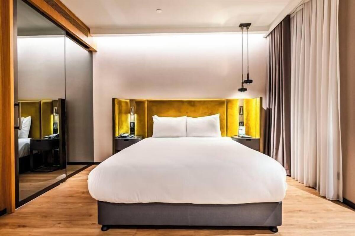 A modern hotel room with a large bed, a wooden floor, a mirror, and a nightstand with a lamp.