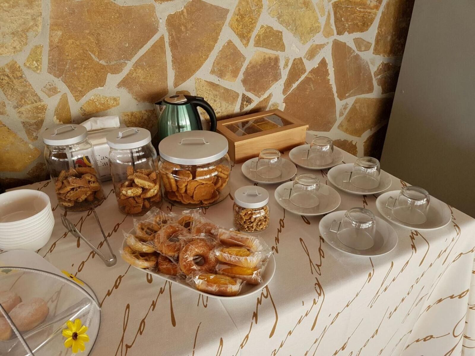 Buffet de petit esmorzar amb diversos aliments, com ara pastissos i cereals, disposats sobre una taula amb un mur de pedra de fons.