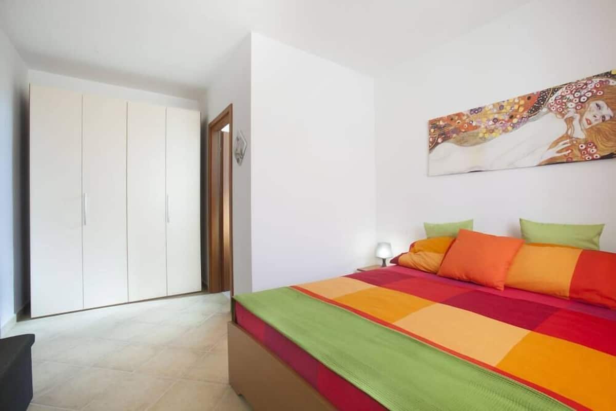 Ein Schlafzimmer mit Bett, farbenfroher Bettwäsche, einem Kleiderschrank und einem Bild an der Wand.