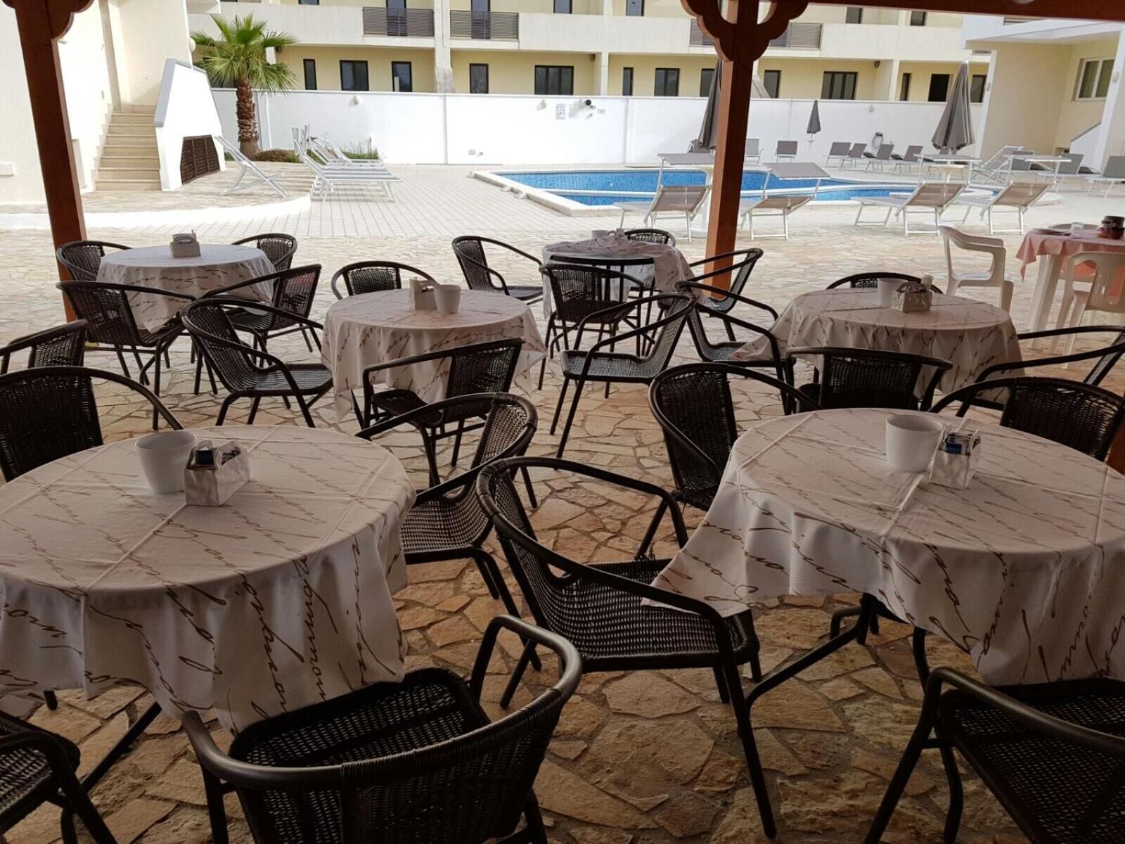 A poolside area with round tables and chairs, a pool, and a building in the background.
