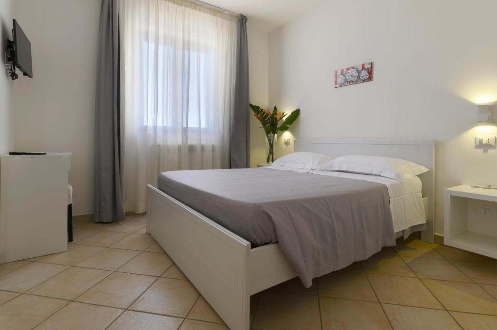 A hotel room with a bed, a bedside table, a TV, a window with curtains, and a wall-mounted light.