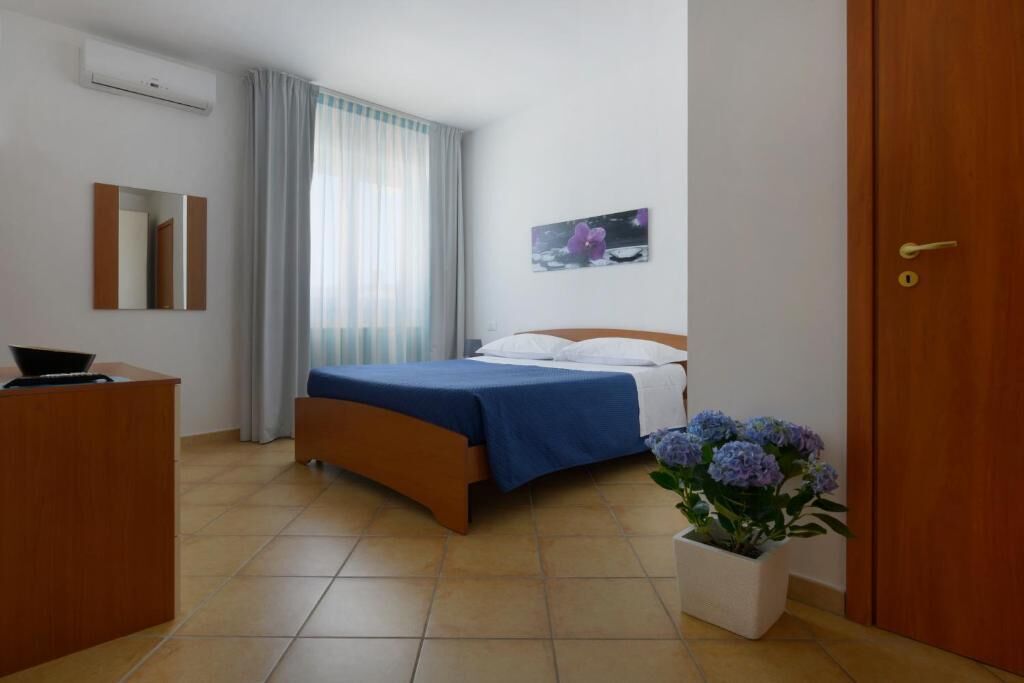 A hotel room with a bed, a desk, a mirror, a window with curtains, a wall-mounted picture, and a potted plant.