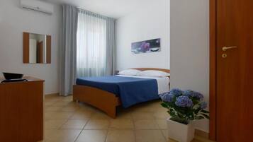 A hotel room with a bed, a desk, a mirror, a window with curtains, a wall-mounted picture, and a potted plant.