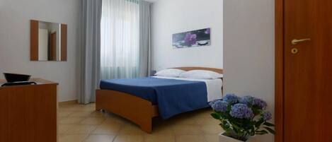 A hotel room with a bed, a desk, a mirror, a window with curtains, a wall-mounted picture, and a potted plant.