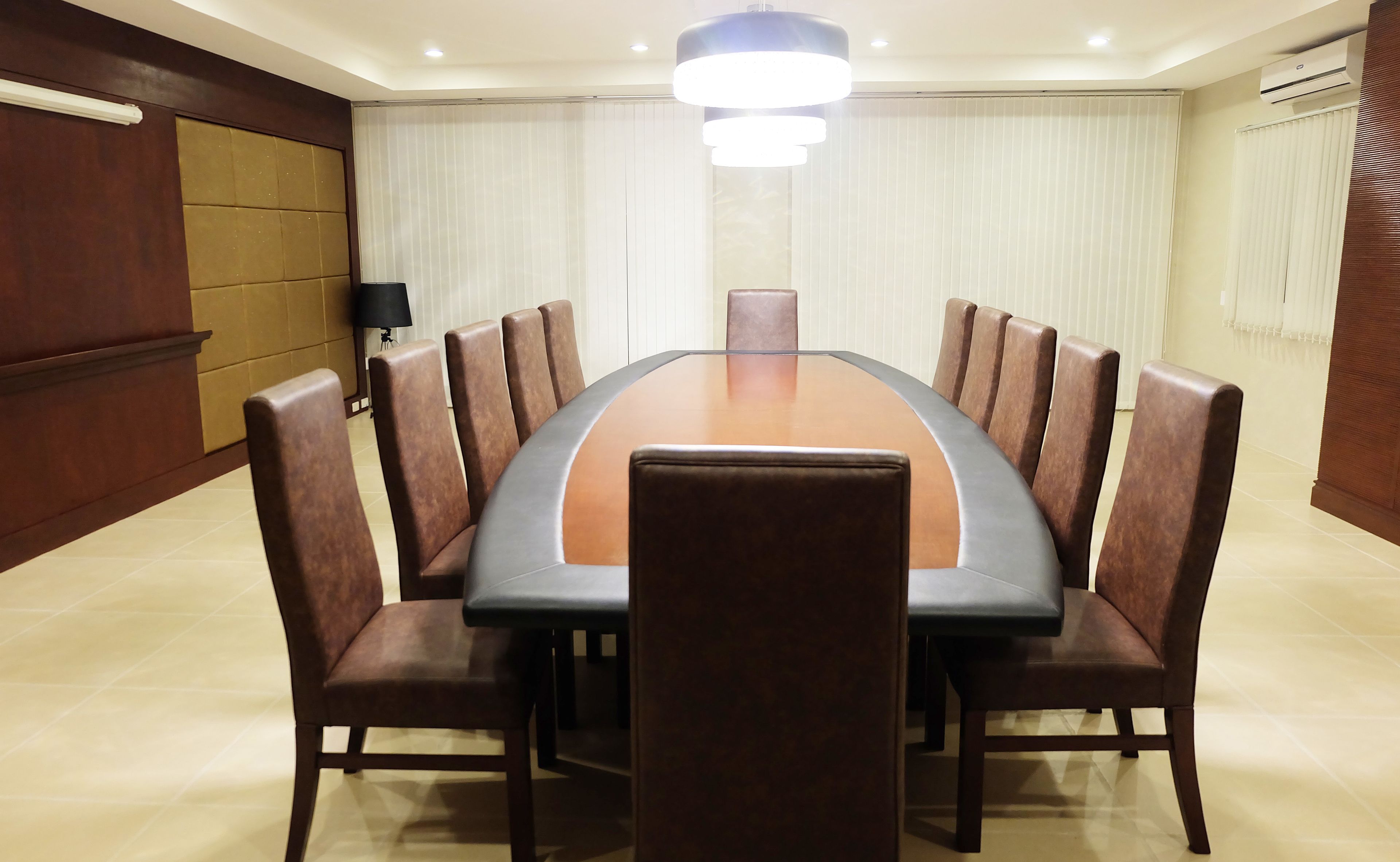 A conference room with a large oval table, eight upholstered chairs, a ceiling light, and a window with blinds.
