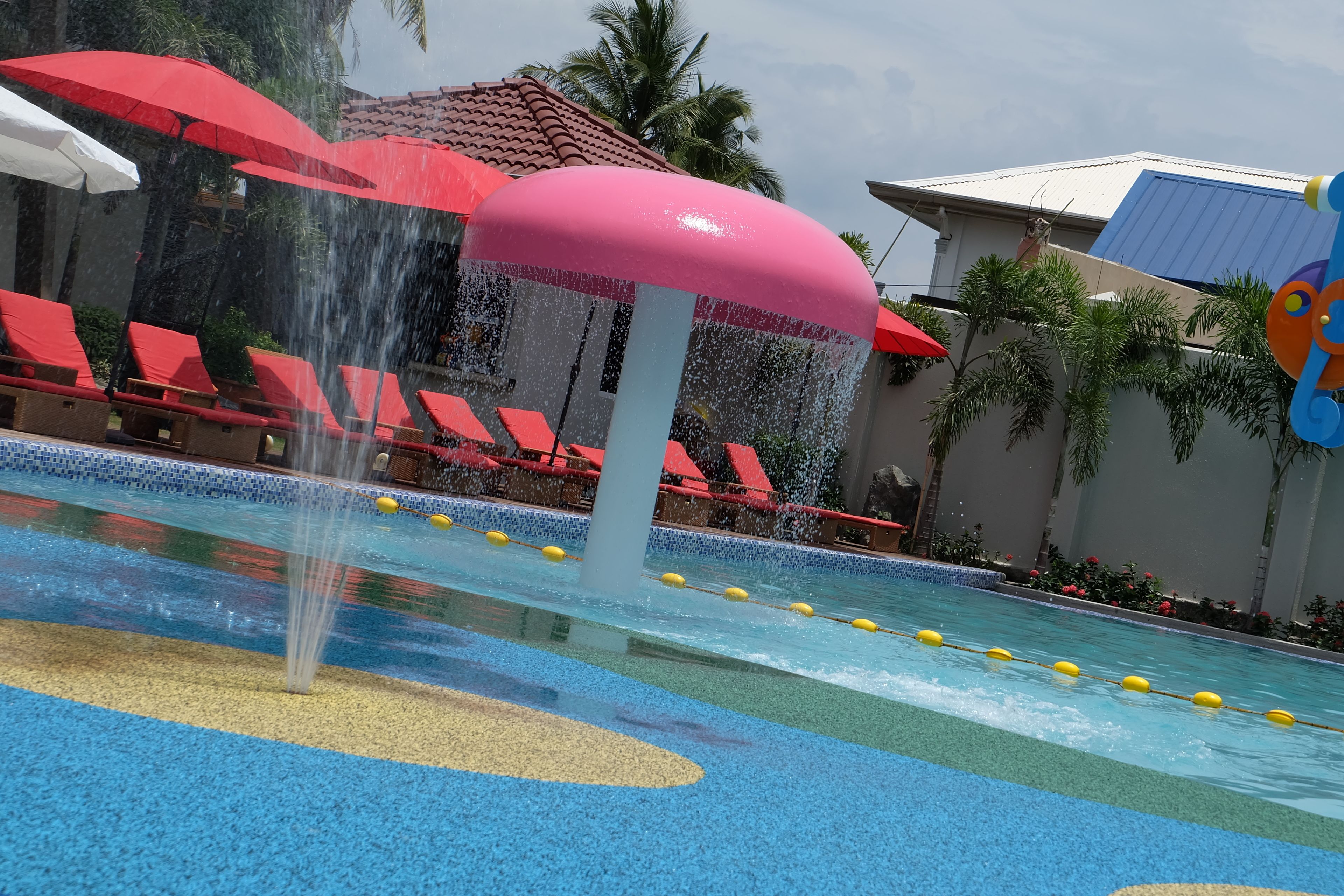 Zona de juegos acuáticos con piscina, sombrillas rojas y sillones de descanso.