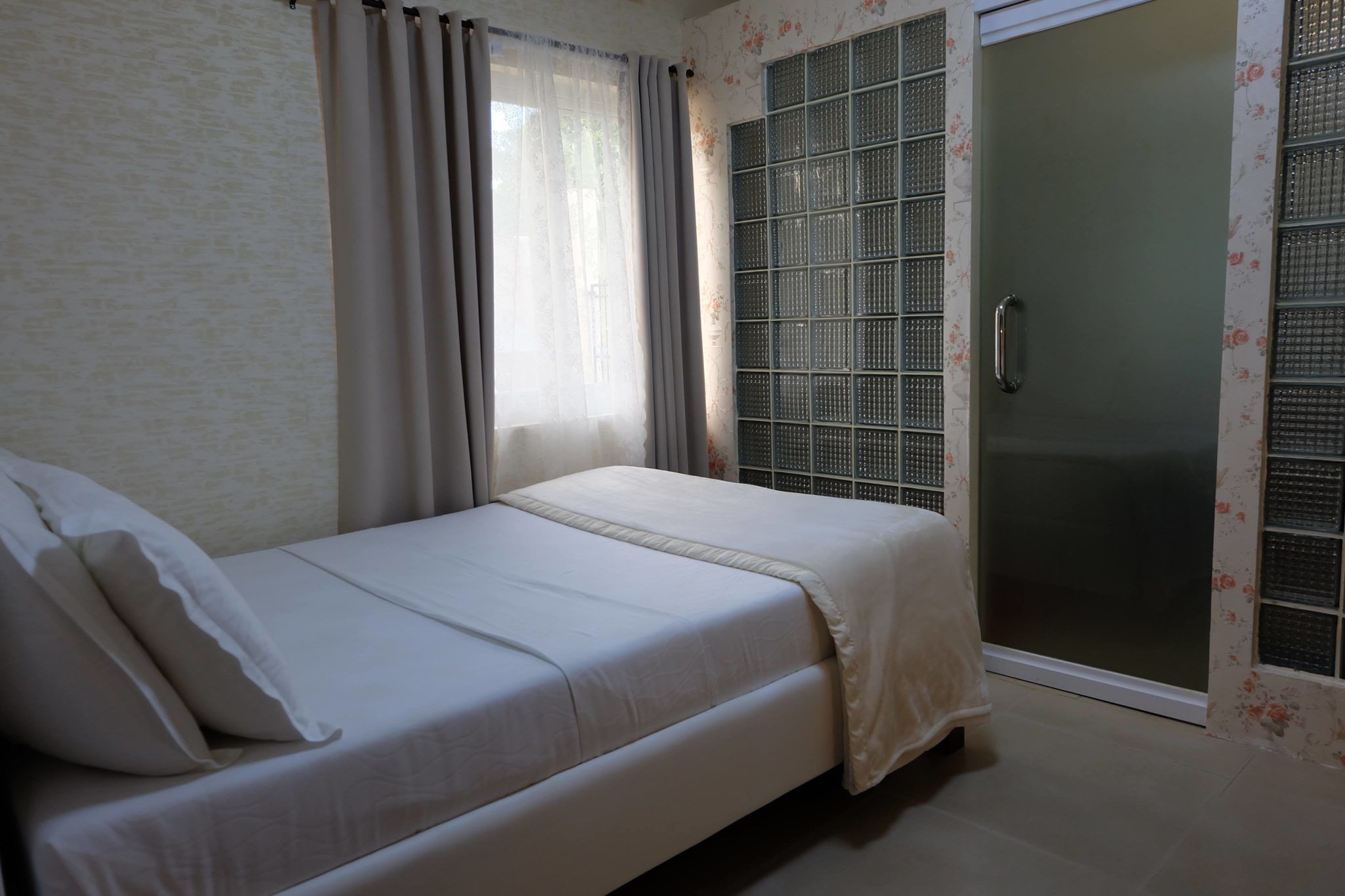 Una cama individual con una colcha y almohadas blancas, una ventana con cortinas traslúcidas y un ventanal corredizo con un diseño de celosía metálica.