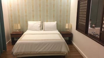 Una cama bien tendida con una colcha blanca y dorada, dos mesitas de noche con lámparas, y una ventana que da a un baño.